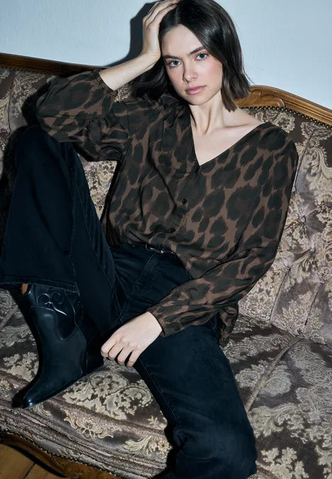 Frau lehnt lässig auf einem gemusterten Sofa, trägt Leo-Bluse, schwarze Jeans und Stiefeletten.